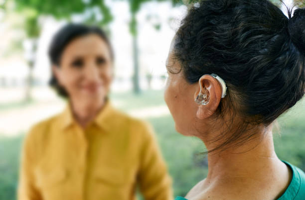 adult woman with a hearing impairment uses a hearing aid to communicate with her female friend at city park. hearing solutions - hearing aid 個照片及圖片檔