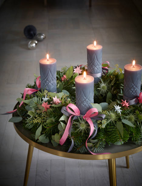 Advent Christmas wreath on brass table. Ribbons tied in bows around lit candles, and folded paper stars.Christmas ornaments in background on floor.