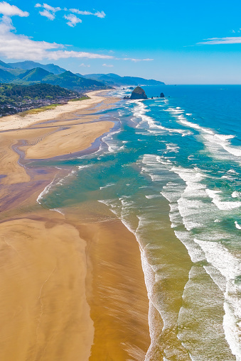 Aerial of Cannon Beach shoreline with a soft mist in the air and warm sandy beaches with blue green water and white foam with Haystack Rock in the distance on a blue sky sunny day with some clouds-color graded