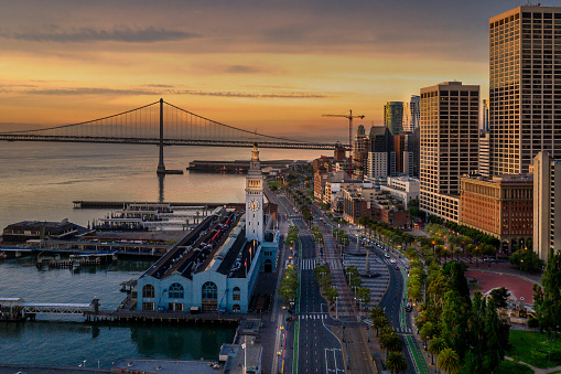 An aerial view of the San Francisco Embarcadero and famous Ferry Terminal at sunrise. Light shines off the glass skyscrapers and the street lights illuminate the street.