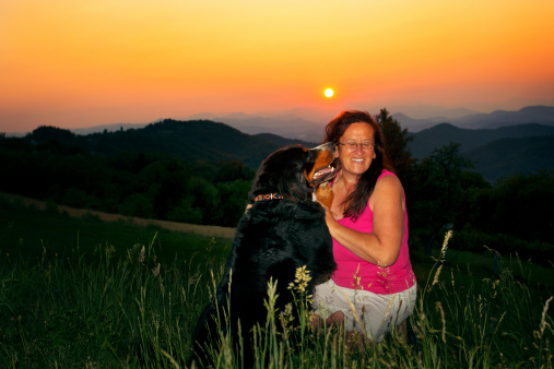 Smilling woman in her 50s and her affectionate dog sharing a happy moment. Sunset in the back.