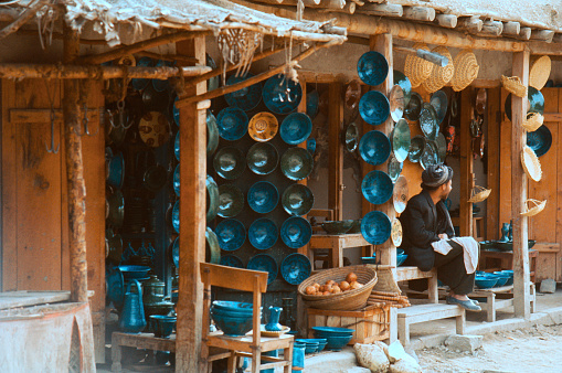 Kabul market in 1969.  An open air stall laid out with deep blue, green and ochre ceramics for sale. Also on display are wicker baskets and a basket of onions.  The stall keeper, not in Afghan dress, but in suit and hat, sits awaiting custom.  Recorded on Ektachrome - the date created is approximate.