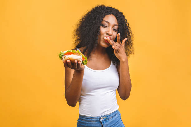 belle jeune femme noire afro-américaine mangeant le hamburger isolé sur le fond jaune. - eating burger photos et images de collection