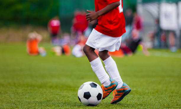 african american boy i junior football team ledande boll på gräs utbildning field. ungdom fotbollsspelare sparkar boll - barndom bildbanksfoton och bilder