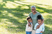 A senior African American woman stands outdoors with her arms around her two grandchildren.  Grandmother is standing in the middle, and the twin boy and girl are each holding her hand.