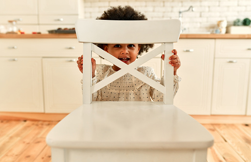 A cute African American little child girl with curly fluffy hair stands behind an armchair and looks out cheerfully smiling in the kitchen at home.