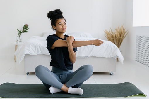 Calm african american girl in sweatpants and t-shirt warming up hands before sport training. Young woman sitting on yoga mat at home, doing stretch exercise. Concept of healthy lifestyle