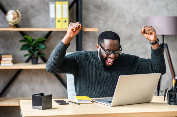 African american guy looks at laptop and rejoices Success at work, good deal. African-American guy looks at the laptop screen, screams excitedly and raises his fists in a victory gesture cheering stock pictures, royalty-free photos & images