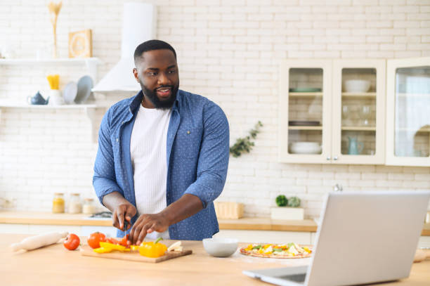 African American guy watching cooking classes Focused handsome African American guy watching cooking classes and learning how to make delicious vegan dinner lunch watching video blog course from laptop in the modern kitchen, chopping veggies cooking class stock pictures, royalty-free photos & images