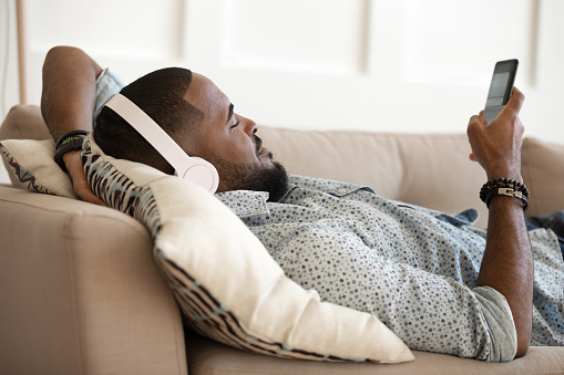 Young handsome african american happy man lying on pillow on comfy sofa, relaxing, listening favorite music in modern wireless bluetooth headphones, chatting in messenger, scrolling social networks.