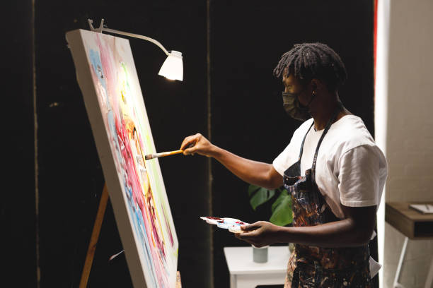 African american male painter wearing face mask painting on canvas in art studio stock photo