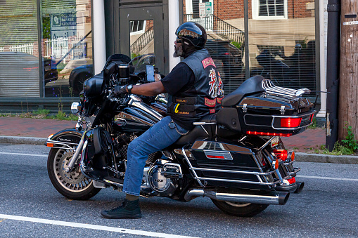 Annapolis, MD, USA 05-02-2021: An African American man with  beard wearing jeans, converse shoes, vest and helmet is idling on the road in the city. He is riding a Harley Davidson motorbike.