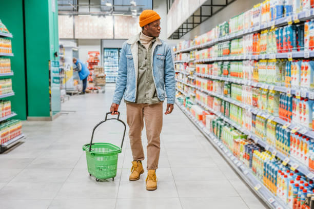 African American man walks with an empty green shopping cart through the supermarket and selects products stock photo
