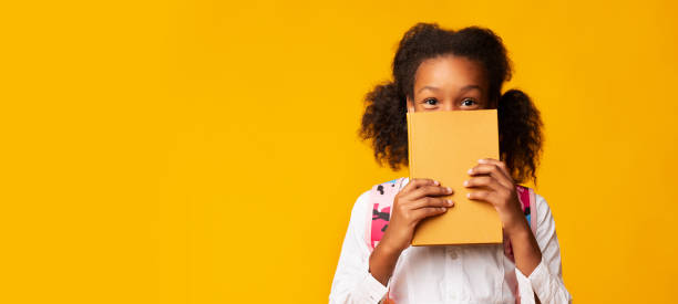 colegiala afroamericana cubriendo la cara con el libro, fondo amarillo - leer fotografías e imágenes de stock