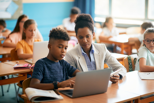 Black female teacher assisting schoolboy in using laptop during a class in the classroom.