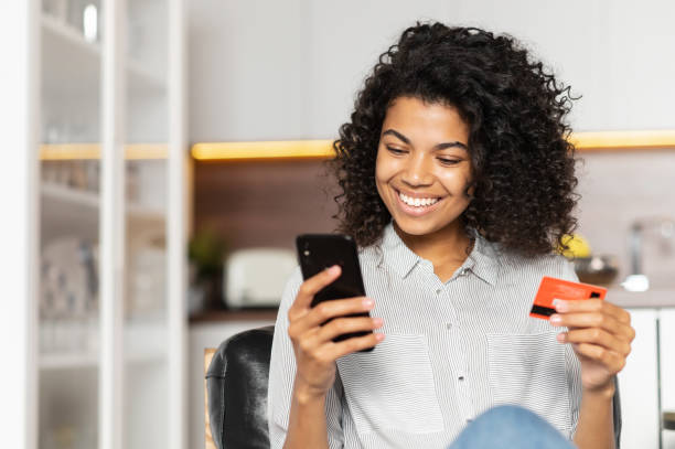African American teenage girl holding credit card Smiling African American teenage girl with curly hair holding mobile phone, entering credit card number to make an online transaction, mixed-race woman ordering food, doing online shopping from home bank account stock pictures, royalty-free photos & images