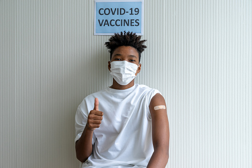 African American teenager giving thumbs-up and showing arm with adhesive patch on arm after received COVID-19 antiviral vaccine to immunization at vaccination station to prevent outbreak coronavirus
