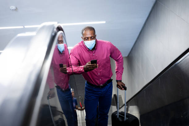 afro-amerikan onun akıllı telefon kullanarak yürüyen merdiven üzerinde duran covid19 coronavirus maskesi giyiyor - south africa covid stok fotoğraflar ve resimler