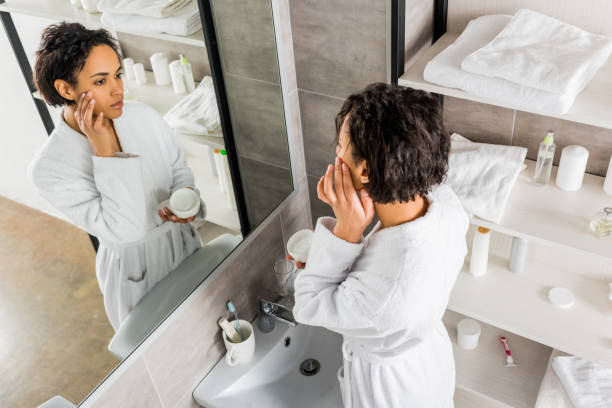 african american woman applying cosmetic anti-wrinkle cream on face and looking in mirror in bathroom african american woman applying cosmetic anti-wrinkle cream on face and looking in mirror in bathroom applying face cream stock pictures, royalty-free photos & images