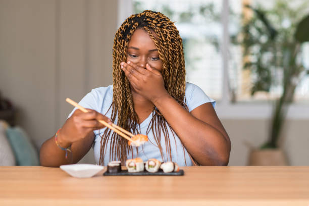 African american woman eating sushi cover mouth with hand shocked with shame for mistake, expression of fear, scared in silence, secret concept African american woman eating sushi cover mouth with hand shocked with shame for mistake, expression of fear, scared in silence, secret concept how do you say shut up in japanese stock pictures, royalty-free photos & images