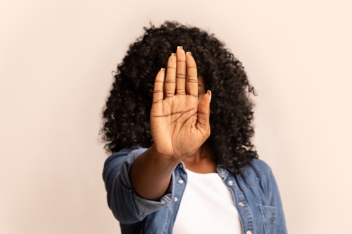 African american curly woman hiding from camera behind her palm over grey studio background. Unrecognizable black lady showing stop gesture, raising one hand, covering face