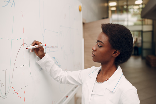 African American woman math teacher writing on blackboard with marker.