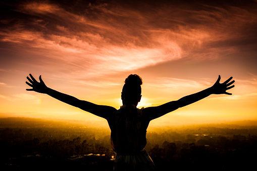 A young African American Woman raises her arms facing the sunset down over the valley and the ocean.