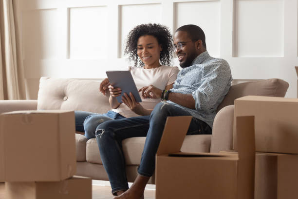 African couple sit on couch near boxes using tablet computer African couple use tablet sit on couch near stack of carton boxes, rest at relocation day, search house renovation ideas, seek shipping company service, shopping online, choose buy things for new home two parents stock pictures, royalty-free photos & images