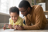 African dad and little creative toddler kid son draw with colored pencils lying on warm floor together, black father baby sitter teaching help child boy learning playing at home, babysitting concept