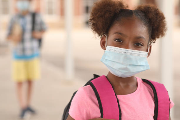 Kembali ke sekolah. Gadis keturunan Afrika di kampus sekolah. Dia memakai masker untuk COVID-19, perlindungan Coronavirus. Siswa lain di latar belakang.