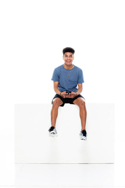 African ethnicity male sitting in front of white background wearing t-shirt and holding box and using mobile phone Full length of aged 18-19 years old with black hair african ethnicity male sitting in front of white background wearing t-shirt who is showing cool attitude and holding box and using mobile phone sitting stock pictures, royalty-free photos & images