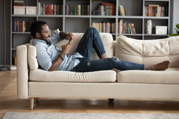 homem africano deita no sofá com websurf de smartphone em casa - video call - fotografias e filmes do acervo