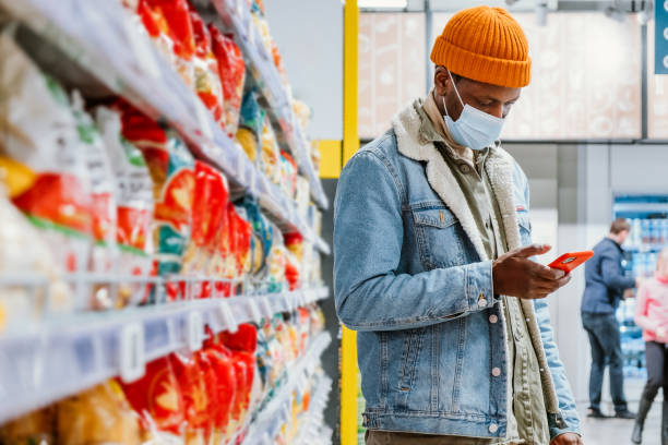 African hipster man wearing a medical mask in a supermarket looks at a list of products on his smartphone while standing at a shelf with goods stock photo