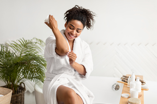Skincare. African American Lady Applying Cream On Dry Elbows Moisturizing Caring For Skin And Body After Morning Shower Sitting On Bathtub In Bathroom At Home. Beauty And Wellness Concept