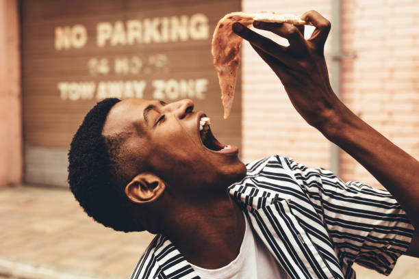 African man eating pizza Young african man eating pizza slice outdoors. Man putting the whole pizza slice in his mouth while sitting outside. mouth open stock pictures, royalty-free photos & images