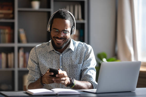 African man holding smartphone wear headphones hearing voice message African man worker or student sit at desk distracted from study or work hold smartphone wear headphones hearing voice audio message from friend, enjoy free time use modern gadget wireless tech concept free images for downloads stock pictures, royalty-free photos & images
