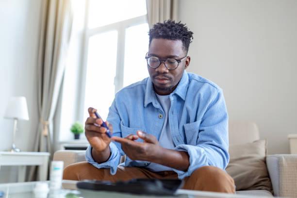 African man is sitting at the sofa and taking blood from his finger due to diabetes. The daily life of a man of African-American ethnicity person with a chronic illness who is using glucose tester. stock photo
