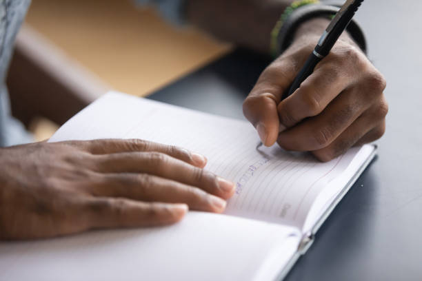 afrikaanse mens die bij bureau wordt gezeten schrijft informatie op notitieboekje close-up - schrijven stockfoto's en -beelden