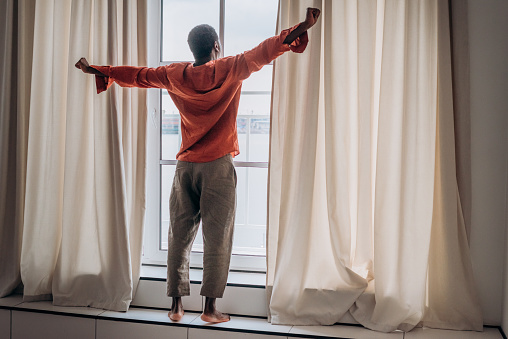 African man in orange pajamas and pants stretches after waking up standing at the window with curtains and looks into the distance, doing morning exercises, the beginning of a new day