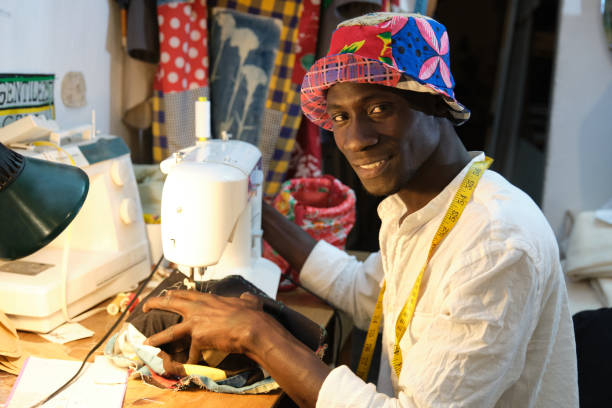 African tailor sewing a hat with sewing machine at sewing workshop smiling and looking at camera. Black man working with african fabrics. stock photo