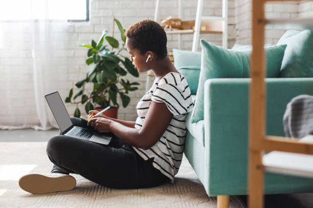 African woman learning online. Standing on the floor listening and noting in the notebook. Online lifestyle stock photo