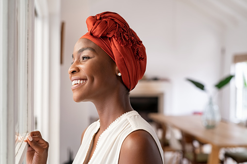 Smiling mid adult african american woman with turban looking outside the window. Cheerful black mature woman wearing traditional red headscarf while contempliting outdoor. Happy middle aged lady looking outside the window at home while thinking.
