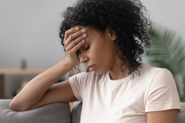 african woman sitting on couch feels unhappy having problems - migraine imagens e fotografias de stock