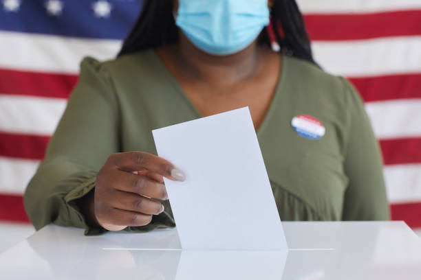 African Woman Voting in Pandemic Close Up Close up of African-American woman wearing mask putting vote bulletin in ballot box and looking at camera while standing against American flag on election day, copy space polling place stock pictures, royalty-free photos & images