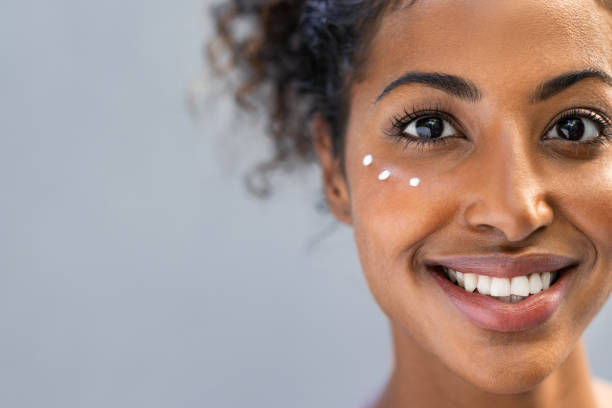 African woman with lotion under the eyes Young african woman applying moisturizer on face isolated on grey background with copy space. Closeup of beautiful girl applying cosmetic cream on skin near eyes and looking at camera. Portrait of smiling african american lady with three dots of cream on cheek. applying face cream stock pictures, royalty-free photos & images