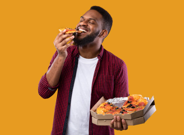 african young man disfrutando de pizza posando con caja, fondo amarillo - comer fotografías e imágenes de stock