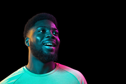 Smiling looking at side. African young man's portrait on dark studio background in neon light. Beautiful male model in casual style. Concept of human emotions, facial expression, youth, sales, ad.