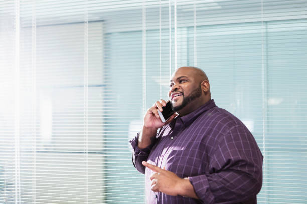 African-American businessman with large build on phone A mature African-American businessman in his 40s standing in an office board room wearing talking on a mobile phone. He is a large man with a shaved head, beard and mustache. He is smiling, looking upward. fat man looks at the phone stock pictures, royalty-free photos & images