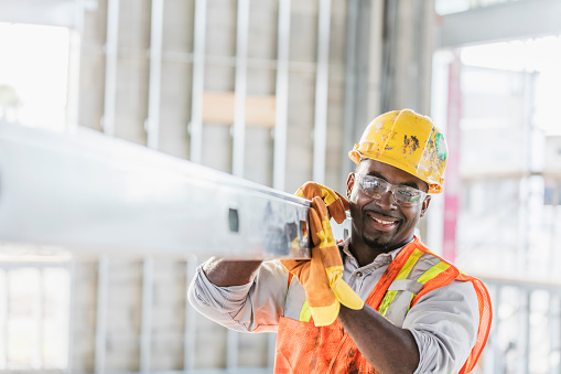A mid adult African-American man in his 30s wearing a hard hat, protective goggles, a safety vest and work gloves, carrying a piece of construction material into the structure being built. The construction worker is smiling, looking at the camera.
