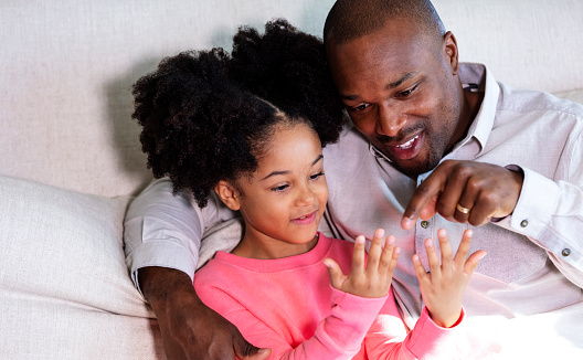An African-American father and daughter sitting side by side together at home on the living room sofa. Dad's arm is around the girl's shoulder. She is holding up her hands and dad is counting the fingers.